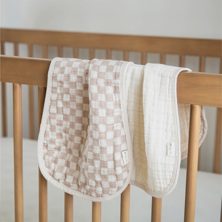 Two Itzy Ritzy muslin burp cloths in checkered mauve and cream draped over wooden crib rail