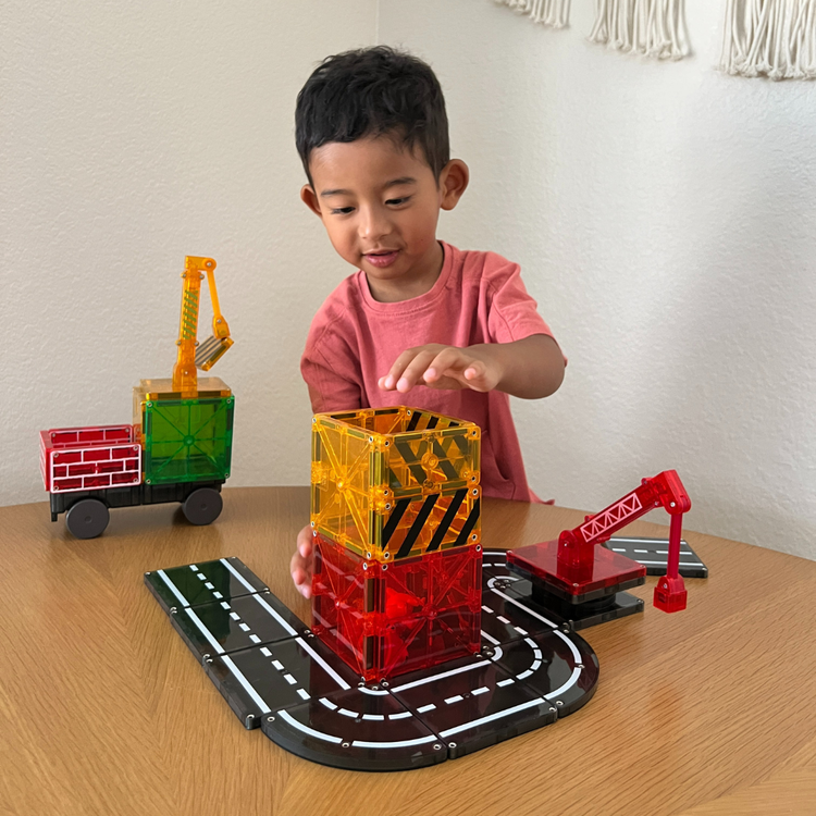 Young child playing with Magna-Tiles Builder 32-piece set featuring translucent magnetic tiles, road tracks, crane, and truck