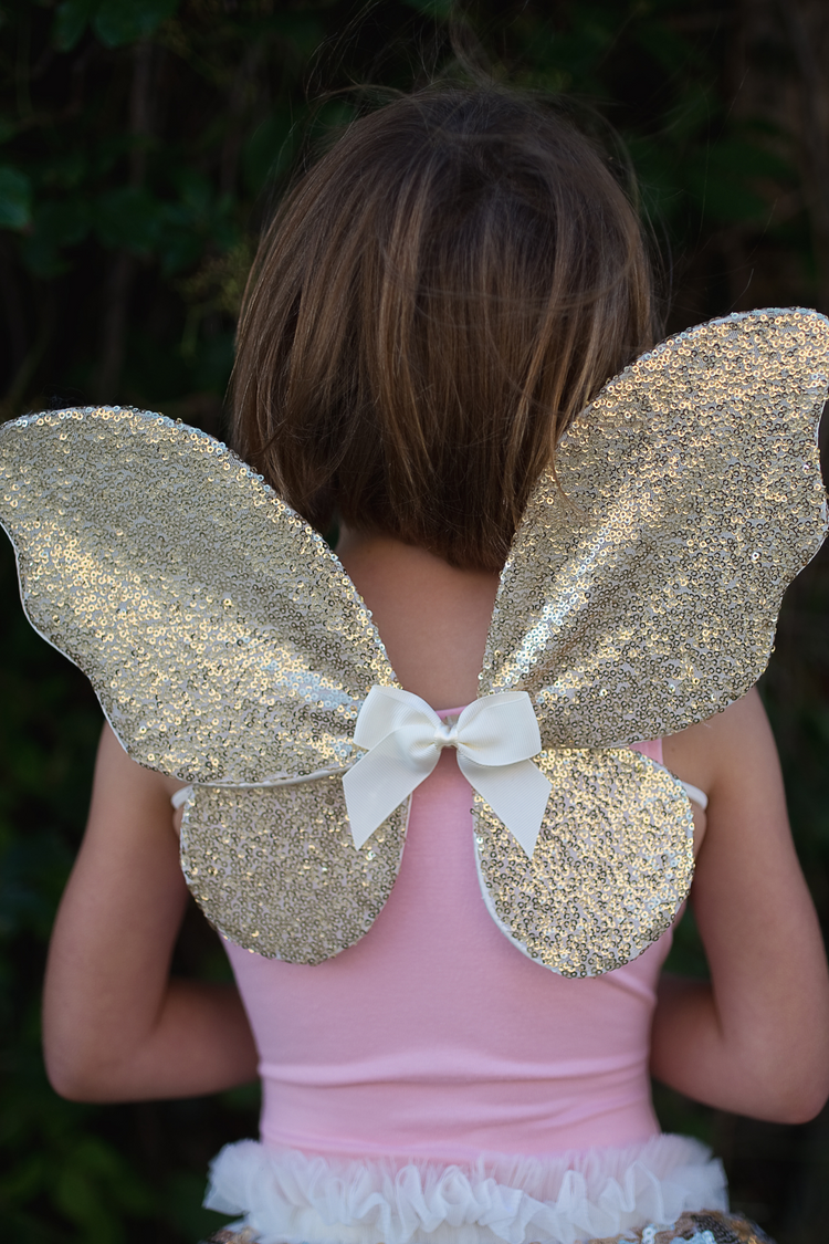 Child wearing gold sequin fairy wings with white bow on pink costume dress