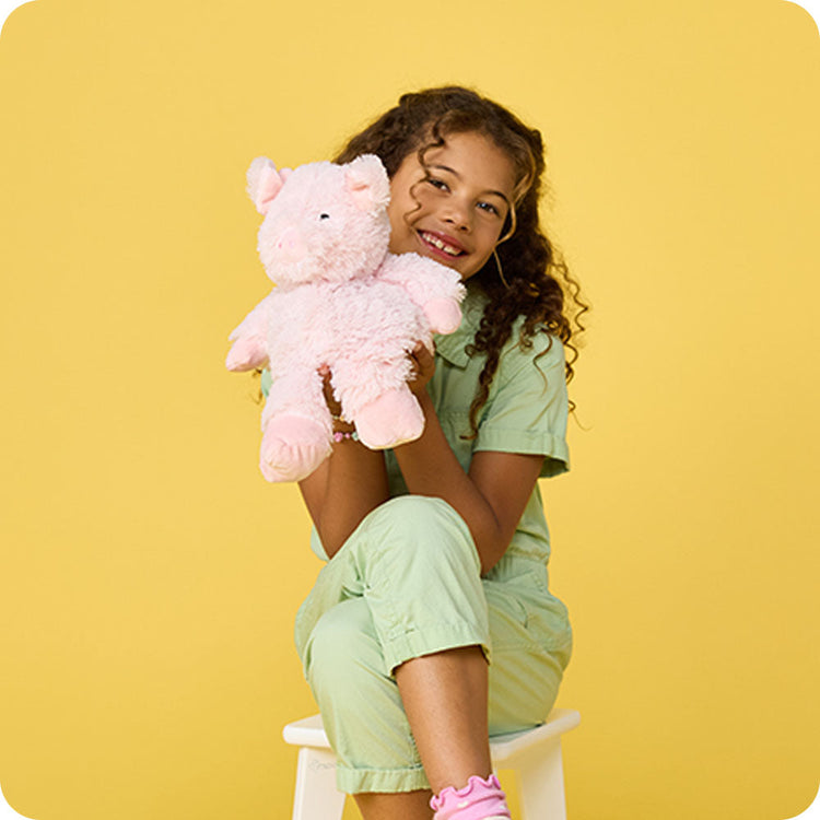 Smiling girl in green outfit hugging a pink fluffy pig Warmies stuffed animal