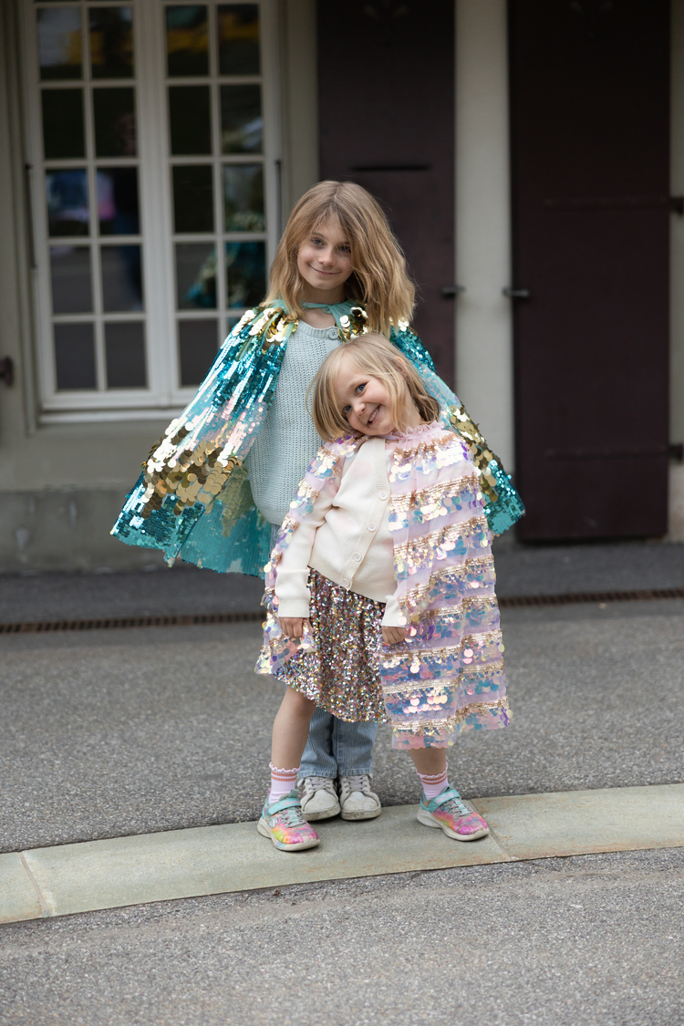 Two girls wearing sequin capes, smaller child in gold light pink pailette cape outdoors