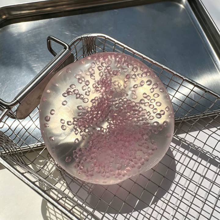 Translucent pearl pink squishy ball with bubble texture resting on metal wire basket