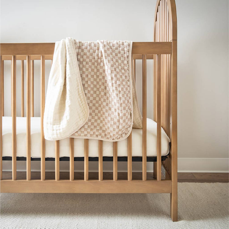 Beige checkered muslin baby blanket draped over a natural wood crib railing