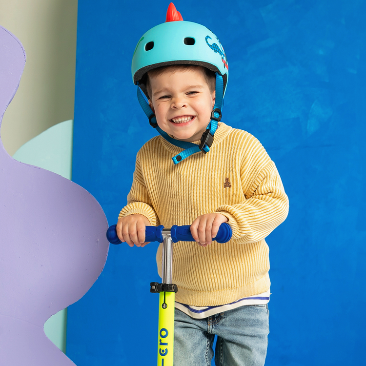 Smiling young child wearing teal dinosaur helmet riding neon yellow Micro Mini scooter with blue handles