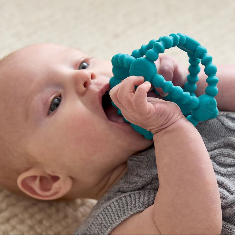 Baby lying down chewing on teal Itzy Ritzy Bitzy Ball silicone teether toy