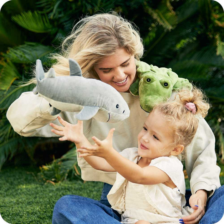 Woman and young girl playing with gray shark and green turtle Warmies Junior plush toys outdoors
