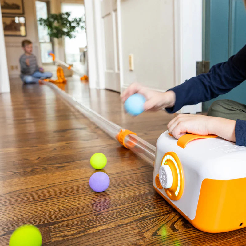 Two children playing with Air Toobz ball launcher and clear tube track indoors on hardwood floor.