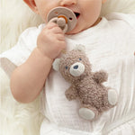 Baby holding a taupe plush bear toy attached to a natural rubber pacifier while lying down