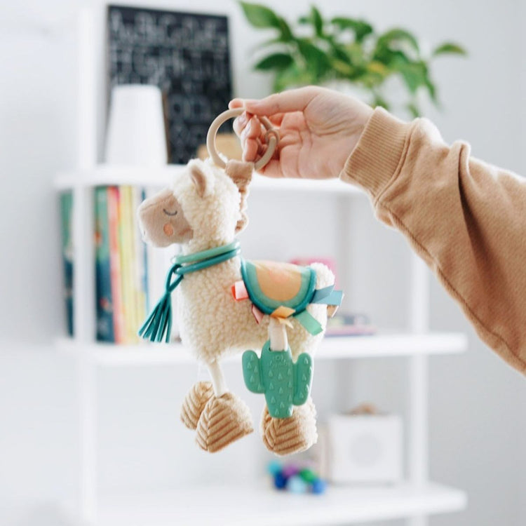 Hand holding Itzy Ritzy llama plush activity toy with teal cactus teether and ring clip