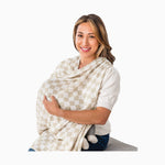 Smiling woman wearing taupe checkerboard nursing cover while breastfeeding on white background