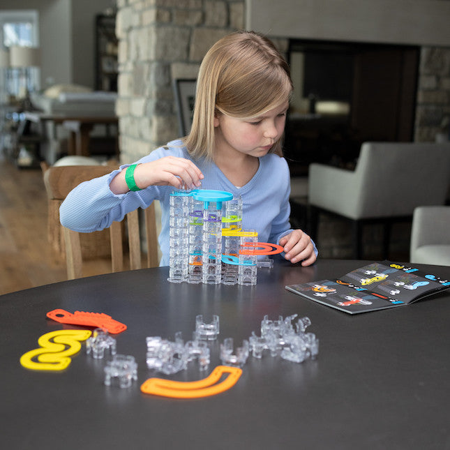 Young girl building a colorful Trestle Track Pro tower with clear interlocking blocks at a table.