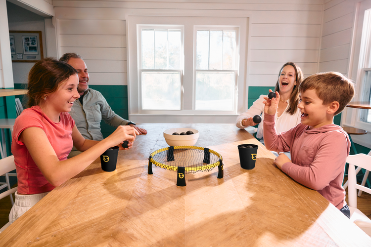 Family of four laughing and playing Spikepong mini game at a wooden dining table indoors.