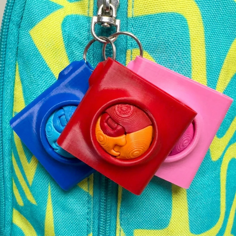 Three colorful Zorble Pod keychain fidget holders in blue, red, and pink attached to a teal backpack zipper.